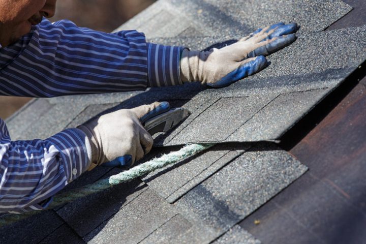 A worker wearing protective gloves using a cutter to make the shingles fit into place